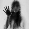 black and white photo of woman behind frosted glass with most of face blurred and hand raised to touch glass