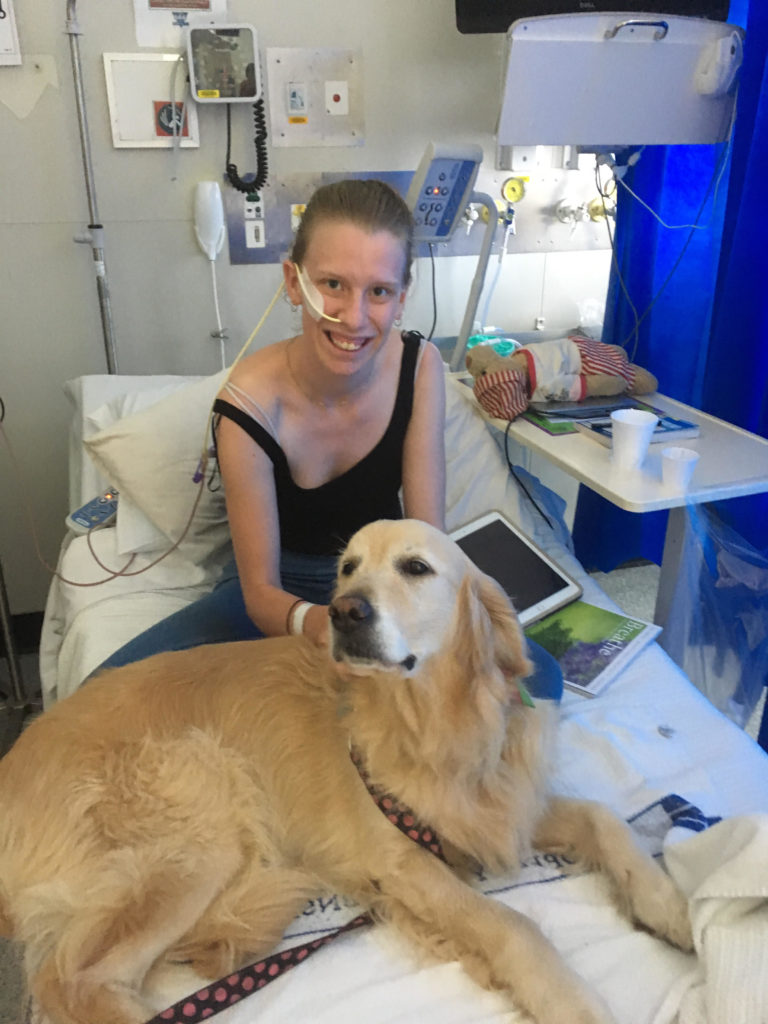 photo of contributor, a young woman on a hospital bed with a feeding tube through her nose and a large golden labrador dog on the bed with her