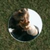 photo of circular mirror sitting on grass while reflection of young blonde woman reaches down to her reflection in the mirror