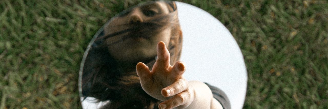 photo of circular mirror sitting on grass while reflection of young blonde woman reaches down to her reflection in the mirror