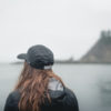 photo of woman wearing baseball cap looking away from camera and staring out at foggy sea horizon