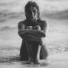 black and white photo of nude woman sitting on beach in waves hugging knees and looking upset into camera