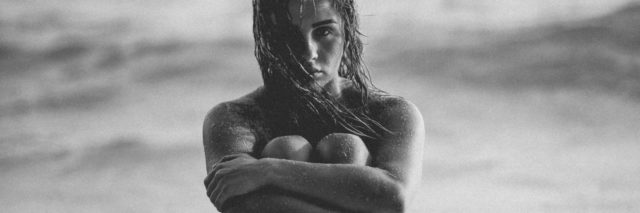 black and white photo of nude woman sitting on beach in waves hugging knees and looking upset into camera