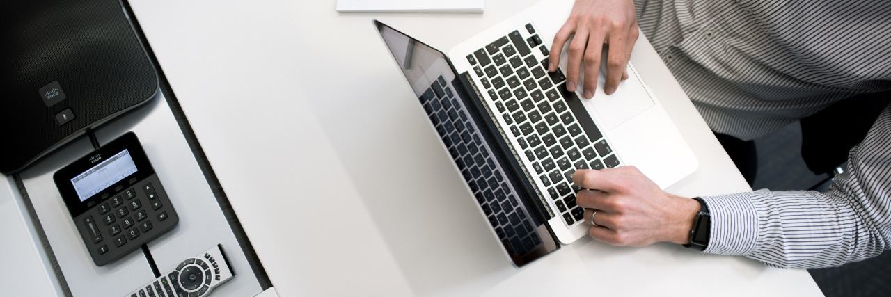 7 Companies With Virtual Jobs Available This May White desk with a phone, tablet, cup of coffee and open laptop with white hands on the keyboard