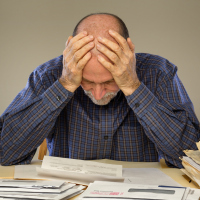 A senior adult man sitting at a table or desk stacked with papers and envelopes looking down with his hands on his head.