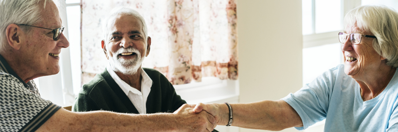 Integrating Into an Assisted Living Community Is a Common Challenge A smiling group of older adults sit around a small dining table with tea. Two men watch as a third person reaches across the table to shake someone’s hand.
