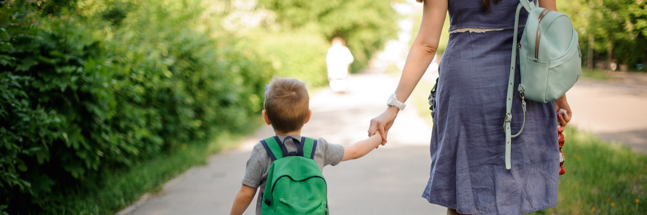 A woman walking with her son, who's wearing a backpack. They are holding hands.