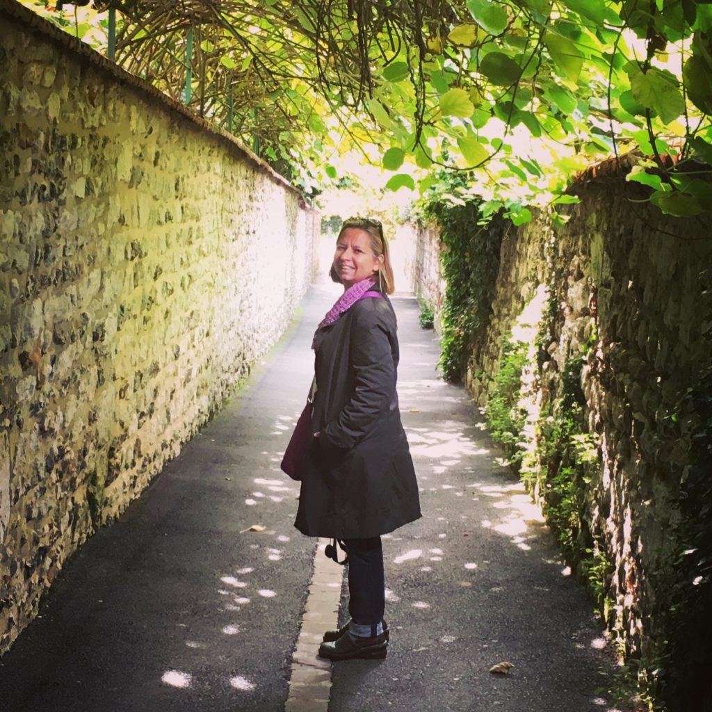 woman standing in an outdoor corridor with stone walls and a ceiling covered with leaves