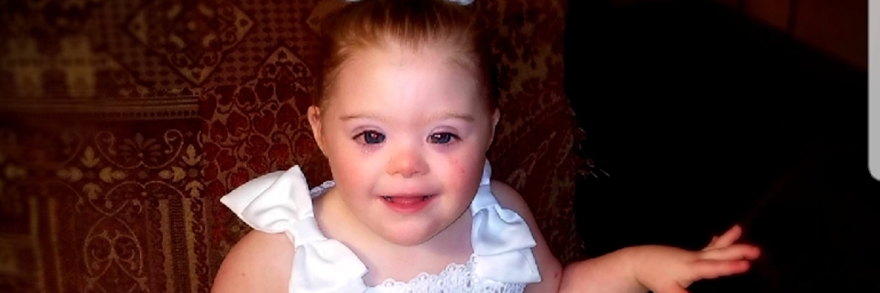 Little girl with Down syndrome sitting on chair, wearing a puffy white dress and smiling at the camera.