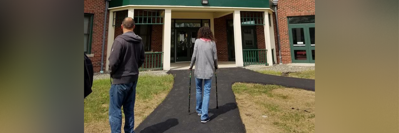 Why I Gave Up Looking for a Cure for My Illness woman walking on asphalt path with her walker toward a brick building