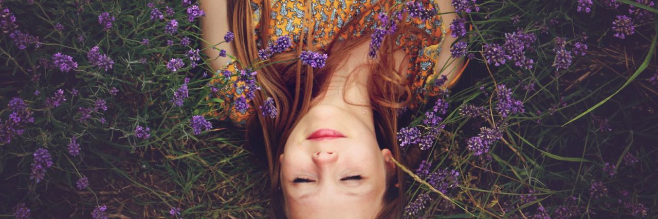 overhead photo of woman lying in purple flowers with eyes closed and smiling