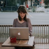 photo of woman writing on laptop outside coffee shop with road behind her