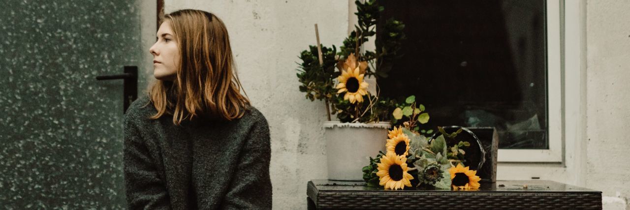 The Loneliness You Experience With Endometriosis photo of young woman sitting outside cottage with sunflowers on small table nearby