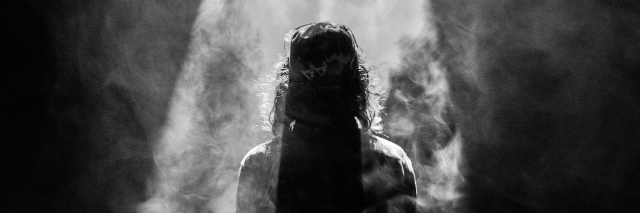 Black and white photo of a person standing in spotlight, surrounded by smoke, and silhouetted by the light.