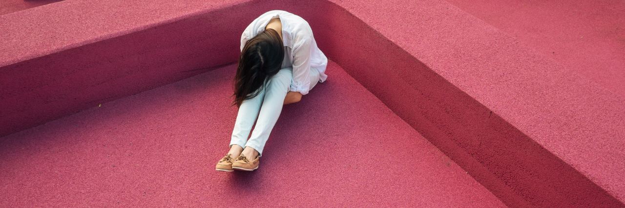 What No One Taught Me About Surviving Rape A woman sitting on a hot pink floor, with her head on her knees looking down