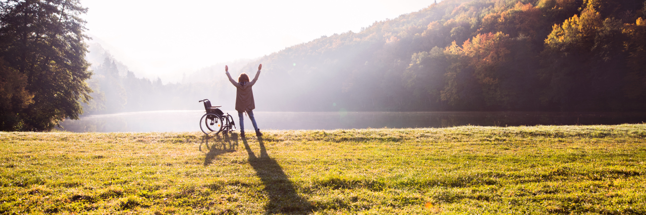 Living With Hypermobile Ehlers-Danlos Syndrome and Using a Wheelchair woman standing with arms in air, next to her wheelchair out in a grassy field