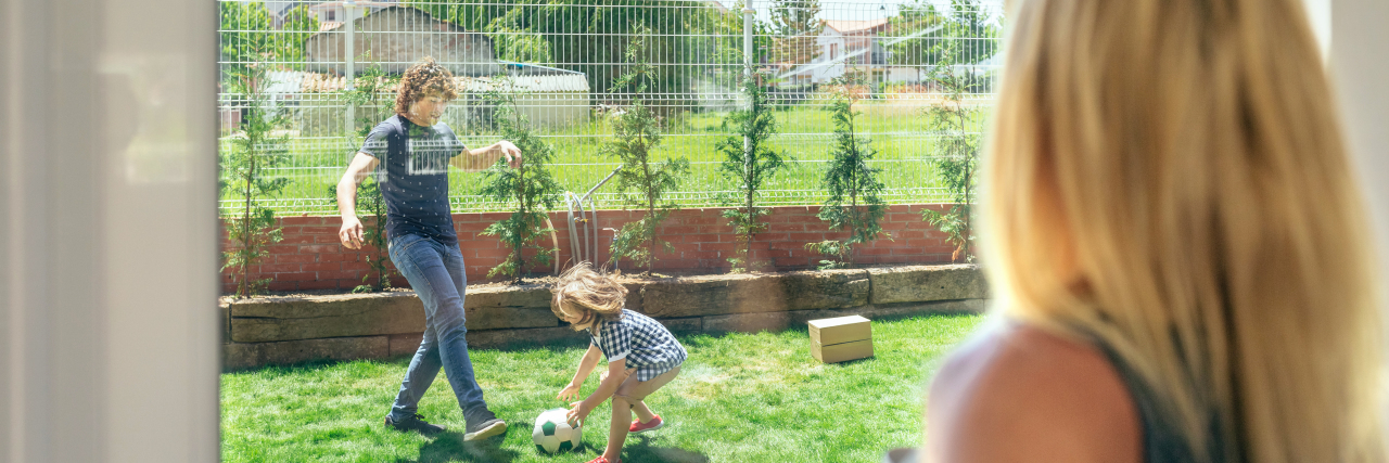 The Emotion That Haunts Me as a Mama With a Chronic Illness a mom is watching her husband and son play soccer while she is inside
