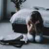 A young girl sitting in front of her bed, holding her head in her hands, looking distressed