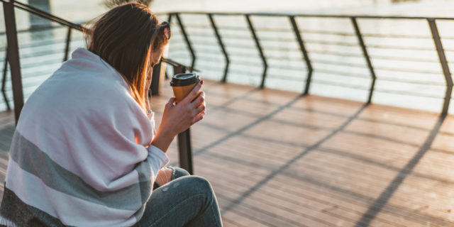 woman sitting with blanket near the water, holding coffee