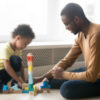father in glasses and adorable toddler son building constructor tower, loving black dad and little child playing with colorful wooden blocks, sitting on warm floor at home