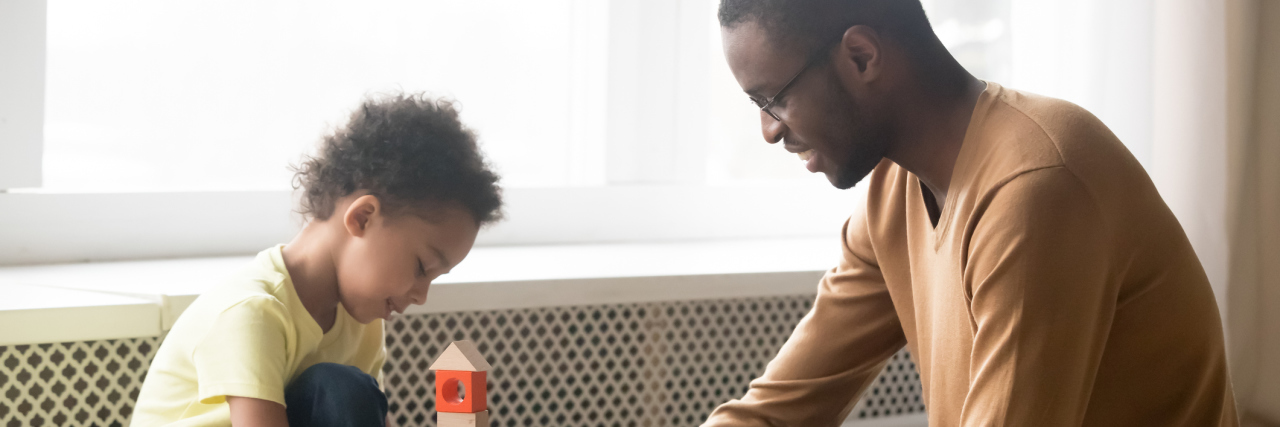 Asperger's Is Now Called Autism - How to Notice ASD in Your Child father in glasses and adorable toddler son building constructor tower, loving black dad and little child playing with colorful wooden blocks, sitting on warm floor at home