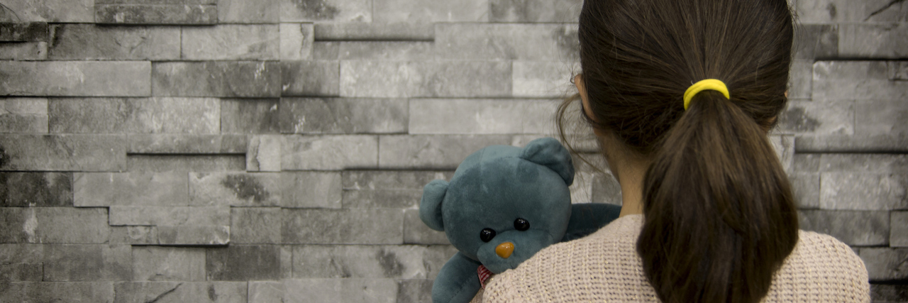little girl holding a teddy near looking at a brick wall