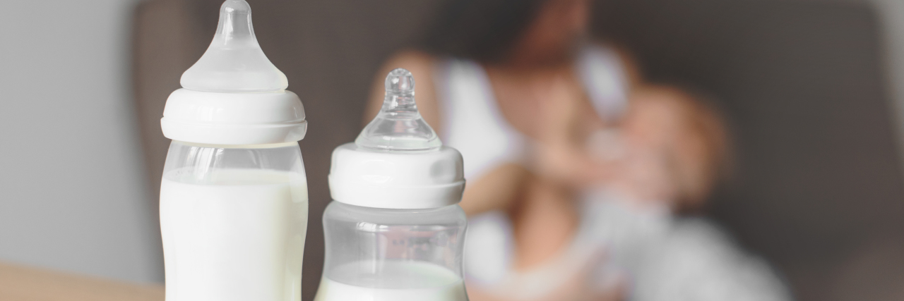 two bottles of breast milk on a table with a mom holding her baby in background