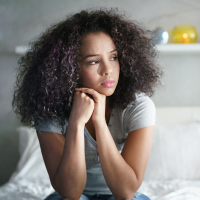 Lonely young woman sitting on bed. Depressed girl at home, looking away with sad expression.