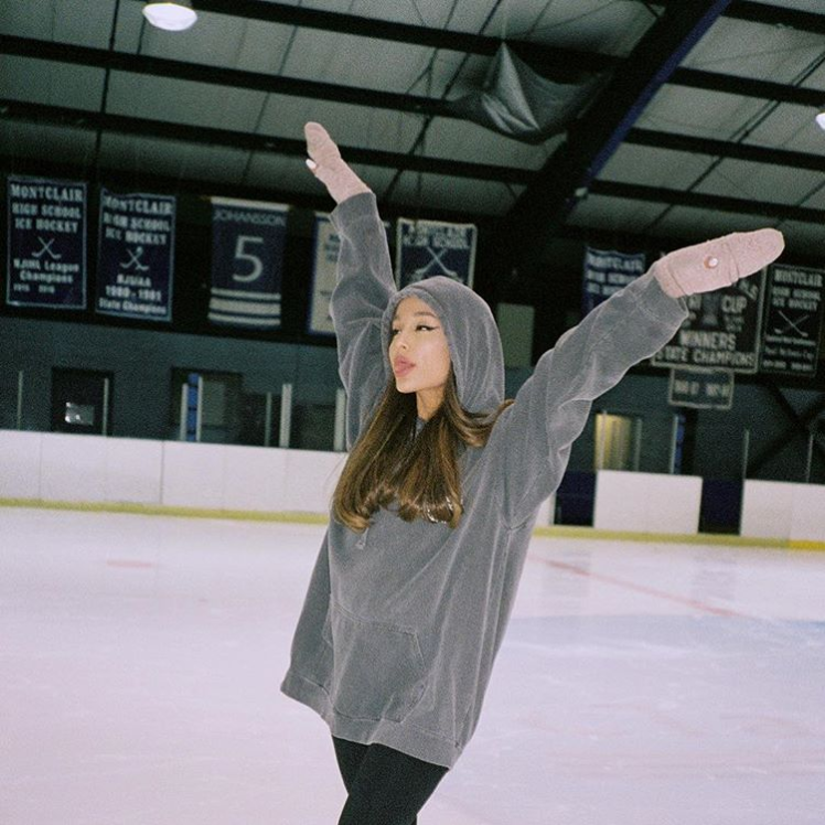 Ariana Grande, singer and actress, throws her hands up in a pose. She is standing in an ice rink. Her hair is long and brown. She is wearing pink mittens and a gray sweatshirt with the hoodie pulled up.
