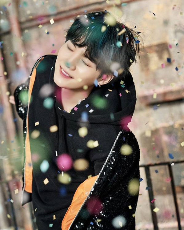 Suga, Korean singer from band BTS, closes his eyes and smiles as confetti rains down. He has black hair and is wearing a black hoodie and sweatshirt.