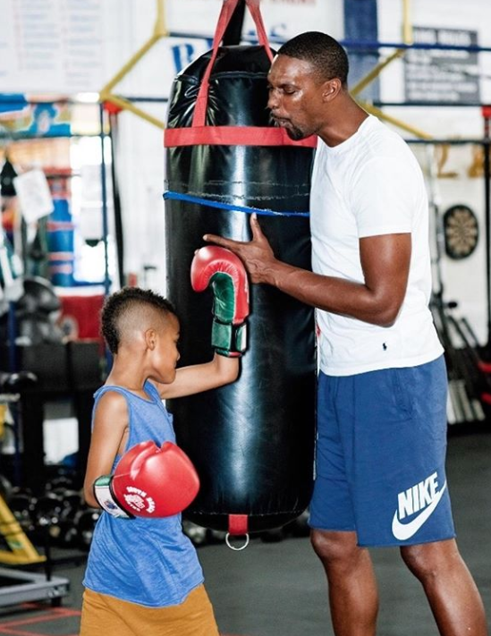 Chris Bosh, African-American NBA player, braces a punching bag while his young son takes a swing. Bosh is wearing a white shirt and blue basketball shorts. His son is wearing red boxing gloves, a blue shirt, and yellow shorts.