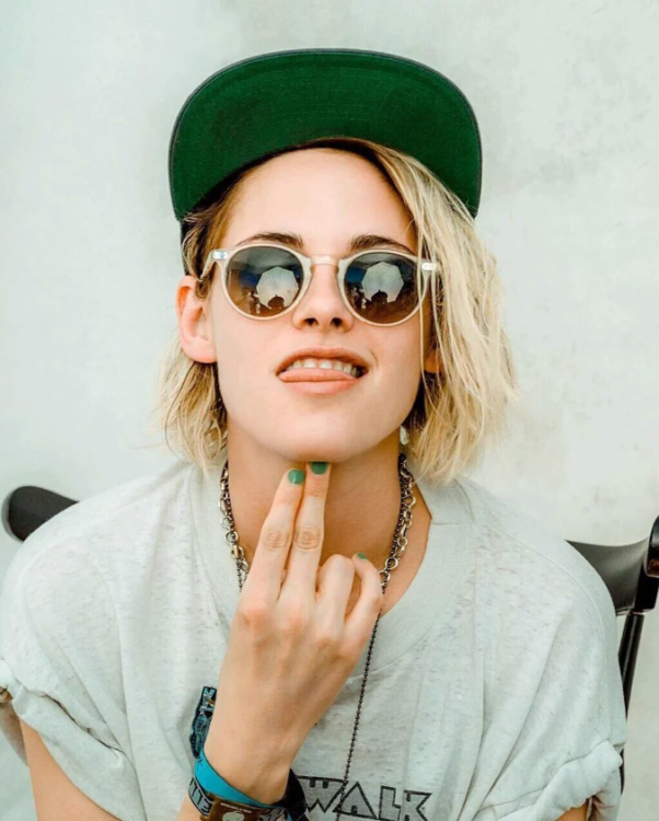 Kristen Stewart, actress, is wearing sunglasses. She has cropped blond hair and is wearing a baseball cap and gray T-shirt. She has placed two fingers on her chin. She is wearing a necklace.