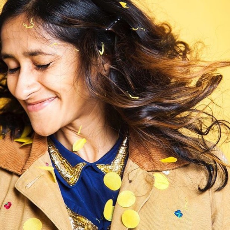 Aparna Nancherla, American comedian, smiles as yellow confetti rains down on her. She has long hair and is wearing a blue blouse and tan blazer.