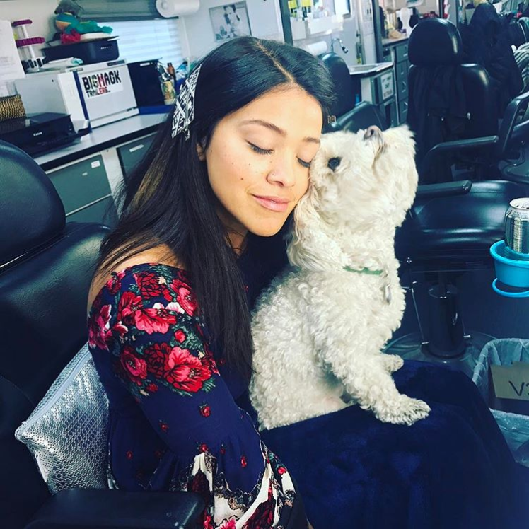 Gina Rodriguez, Latin-American actress, hugs her dog. She is wearing a black shirt with pink and red embroidered flowers. She has long, dark brown hair. Her dog is small and has curly white fur.