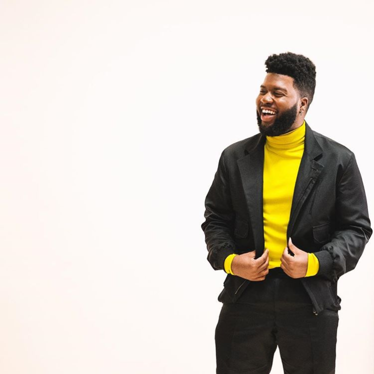 Khalid, American singer, poses in front of an off-white background. He has black hair and a beard. He is wearing a yellow turtleneck and a black suit. He is smiling.
