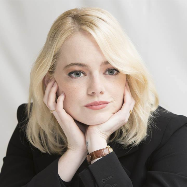 Emma Stone, actress, is cupping her chin in her hands and smiling slightly. She has light blond hair and is wearing a black suit and a watch.