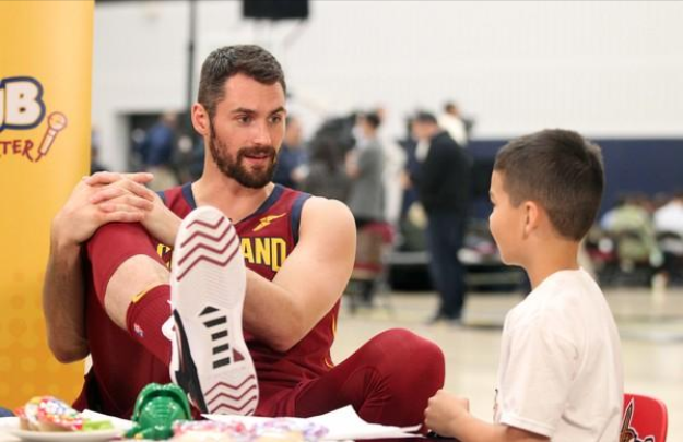 Kevin Love, NBA player, sits and talks to a young boy. He is wearing a red Cleveland Cavs basketball uniform with blue and gold trim. He has short brown hair and a beard. The boy is wearing a white T-shirt.