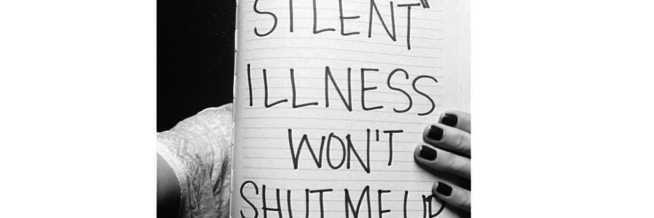 Why I Speak Out About My Chronic Illness black and white photo of a woman holding up a sign that says "my 'silent' illness won't shut me up"