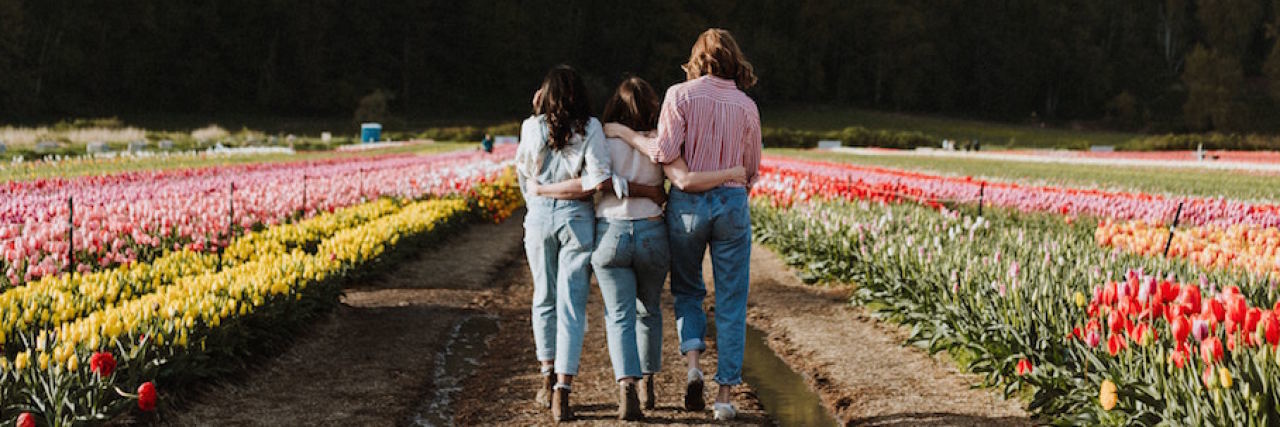 The Types of Friends That Can Help Your Mental Health Three friends with their hands arm and arm, walking through a field of flowers. They're facing away