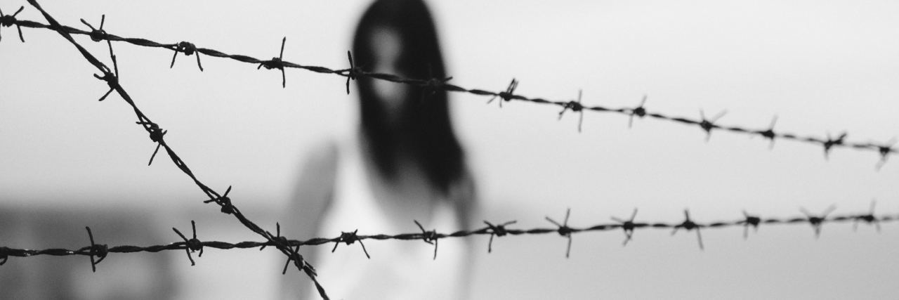 black and white photo of woman in white dress and dark hair, blurred, with barbed wire in foreground