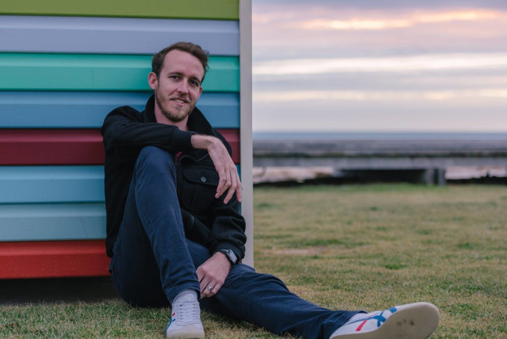 the author sitting outside against a multicolored building with a sunset in the background. he's resting one arm on his knee and smiling