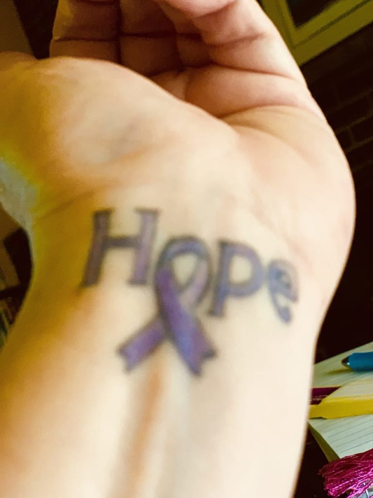 tattoo on a woman's wrist that says "hope." the "o" is the loop in a purple awareness ribbon representing migraine.