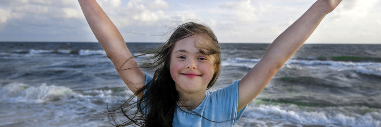 Girl with Down syndrome smiling with arms spread wide and ocean in the background