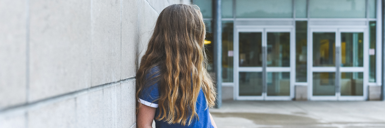 A teenager with long blond hair leaning against a wall, looking behind her.