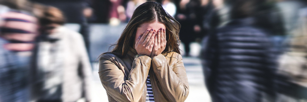 Helping a Friend Having a Panic Attack In Public A woman covering her head with her hands while standing on a crowded street. Everyone else around her is a blur.