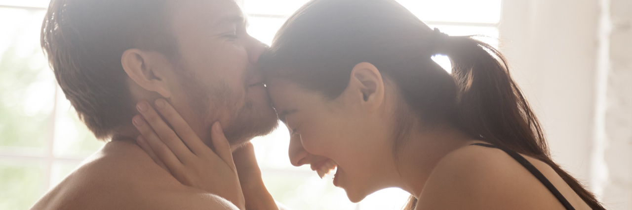 When I Discovered Having Sex Helps My Stutter A man kissing a woman on the forehead as they're sitting in bed.