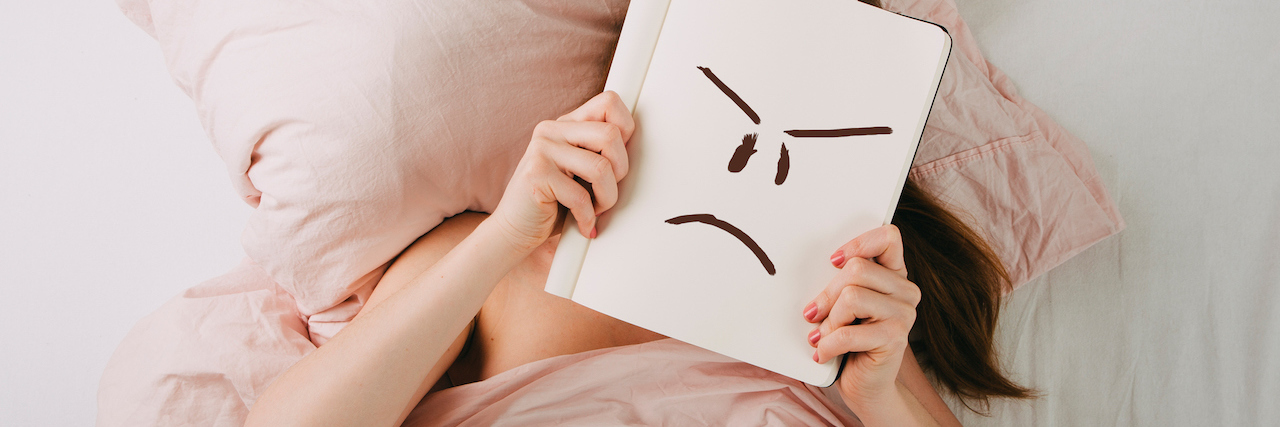 A woman in bed, holding a notebook over her face. On the notebook, there's an anger face drawn in black marker.