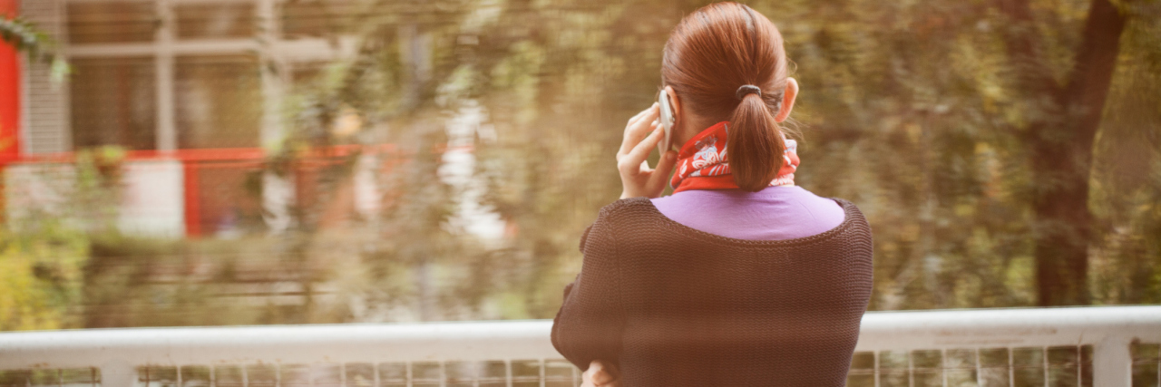 When I Told My Friend Losing Weight Was a 'Silver Lining' to Illness Woman standing on porch in front of her office, using smartphone; candid image shot through window, blurry;
