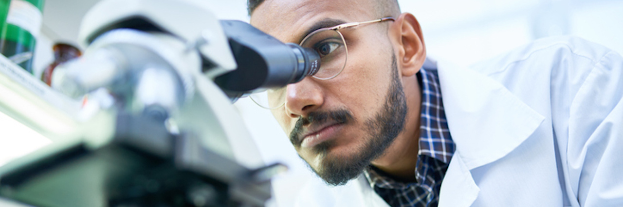 Portrait of young Middle-Eastern scientist looking in microscope while working on medical research in science laboratory, copy space (Portrait of young Middle-Eastern scientist looking in microscope while working on medical research in science laborat