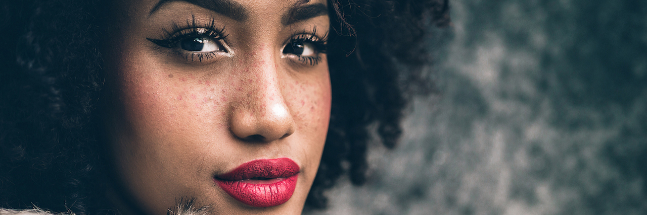 A woman with brown skin and freckles wearing red lipstick and a faux fur coat. Her hair is curly